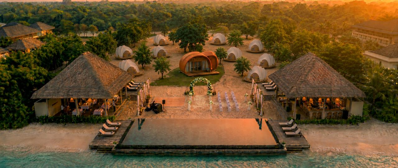 Concept render — the symmetrical beachfront wedding estate at sunset: pool and ceremony deck centred, restaurant and bar pavilions either side, shell tents among the trees
